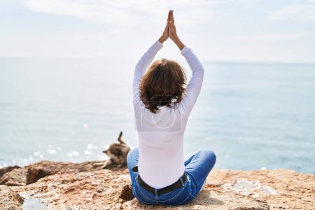 Foto de Mujer de mediana edad haciendo ejercicio de yoga sentado en la roca a la orilla del mar - Imagen libre de derechos