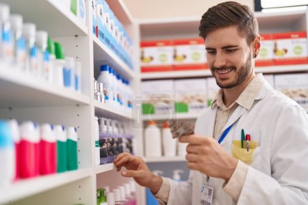 Photo for Young caucasian man pharmacist holding pills of shelving at pharmacy - Royalty Free Image