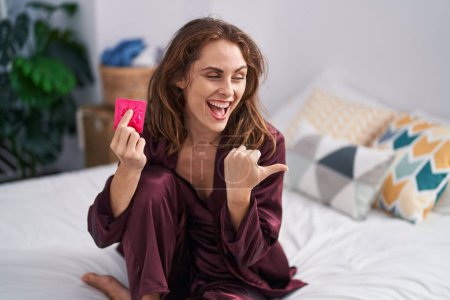 Téléchargez les photos : Belle femme brune tenant un préservatif dans la chambre pointant le pouce vers le côté souriant heureuse avec la bouche ouverte - en image libre de droit