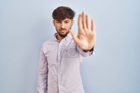Photo for Arab man with beard standing over blue background doing stop sing with palm of the hand. warning expression with negative and serious gesture on the face. - Royalty Free Image