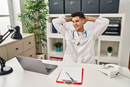 Téléchargez les photos : Young hispanic man wearing doctor uniform relaxed with hands on head at clinic - en image libre de droit