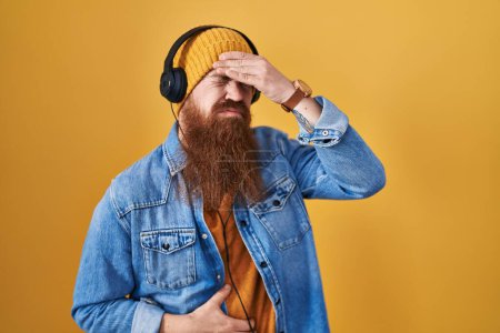 Foto de Hombre caucásico con barba larga escuchando música usando auriculares tocando la frente por enfermedad y fiebre, gripe y resfriado, virus enfermo - Imagen libre de derechos