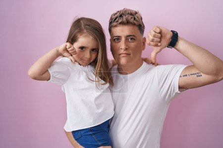 Foto de Padre joven abrazando a la hija sobre fondo rosa con la cara enojada, signo negativo que muestra aversión con los pulgares hacia abajo, concepto de rechazo - Imagen libre de derechos
