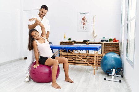 Foto de Hombre y mujer latinos vistiendo uniforme de fisioterapeuta teniendo sesión de rehabilitación de embarazo usando balón en forma en clínica de fisioterapia - Imagen libre de derechos