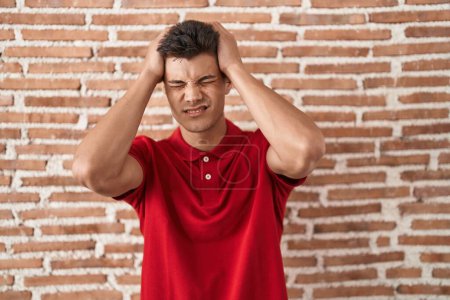 Foto de Young hispanic man standing over bricks wall suffering from headache desperate and stressed because pain and migraine. hands on head. - Imagen libre de derechos