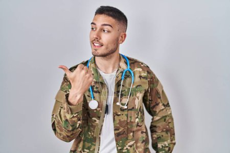 Foto de Young hispanic doctor wearing camouflage army uniform pointing thumb up to the side smiling happy with open mouth - Imagen libre de derechos