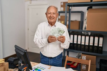 Foto de Hombre mayor con el pelo gris que trabaja en el comercio electrónico de la pequeña empresa sosteniendo dólares buscando de pie positivo y feliz y sonriendo con una sonrisa confiada mostrando los dientes - Imagen libre de derechos