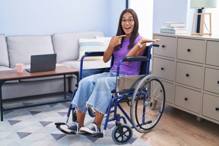 Foto de Joven mujer hispana sentada en silla de ruedas en casa sorprendida y sonriendo a la cámara mientras se presenta con la mano y señalando con el dedo. - Imagen libre de derechos