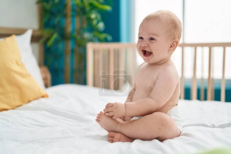 Téléchargez les photos : Adorable bébé caucasien souriant confiant assis sur le lit dans la chambre - en image libre de droit