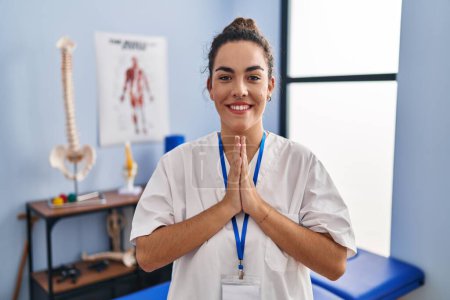 Photo for Young hispanic woman working at rehabilitation clinic praying with hands together asking for forgiveness smiling confident. - Royalty Free Image