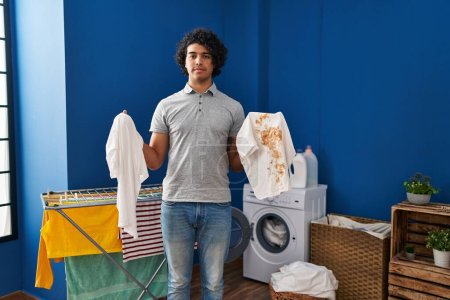 Photo for Hispanic man with curly hair holding clean white t shirt and t shirt with dirty stain relaxed with serious expression on face. simple and natural looking at the camera. - Royalty Free Image