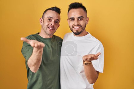 Téléchargez les photos : Couple homosexuel debout sur fond jaune souriant joyeuse présentation et pointant avec la paume de la main en regardant la caméra. - en image libre de droit