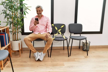 Téléchargez les photos : Moyen-âge homme aux cheveux gris en utilisant smartphone assis sur la chaise dans la salle d'attente - en image libre de droit