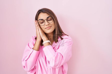 Téléchargez les photos : Young hispanic woman wearing glasses standing over pink background sleeping tired dreaming and posing with hands together while smiling with closed eyes. - en image libre de droit