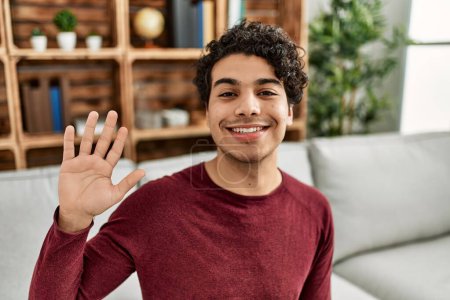 Téléchargez les photos : Jeune homme hispanique souriant heureux de dire bonjour avec la main à la maison. - en image libre de droit