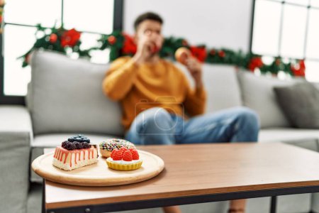 Téléchargez les photos : Jeune homme arabe prenant le petit déjeuner assis sur le canapé par décor de Noël à la maison. - en image libre de droit