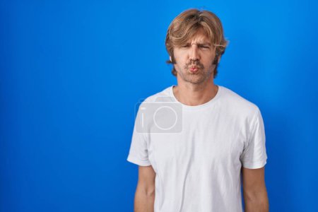 Foto de Middle age man standing over blue background puffing cheeks with funny face. mouth inflated with air, crazy expression. - Imagen libre de derechos