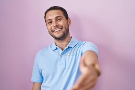 Foto de Hombre hispano de pie sobre fondo rosa sonriente amistoso ofreciendo apretón de manos como saludo y bienvenida. negocios exitosos. - Imagen libre de derechos