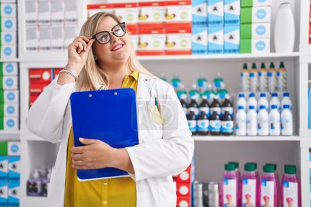Téléchargez les photos : Jeune femme pharmacien souriant confiant tenant presse-papiers à la pharmacie - en image libre de droit