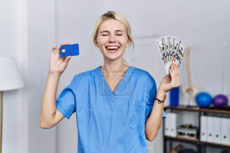 Foto de Joven fisioterapeuta mujer sosteniendo tarjeta de crédito y billetes de dólares sonriendo y riendo en voz alta porque divertido chiste loco. - Imagen libre de derechos