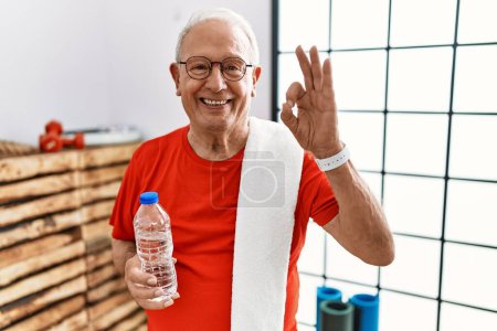 Foto de Hombre mayor con ropa deportiva y toalla en el gimnasio sonriendo positiva haciendo signo de ok con la mano y los dedos. expresión exitosa. - Imagen libre de derechos
