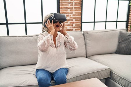 Foto de Mujer de mediana edad jugando a videojuegos con gafas de realidad virtual en casa - Imagen libre de derechos