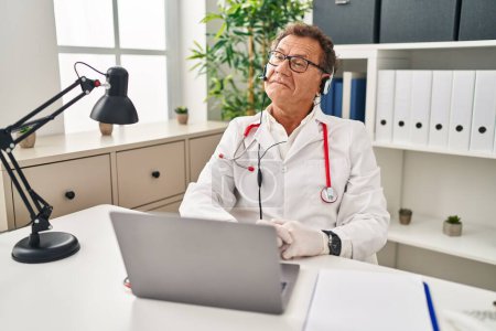 Téléchargez les photos : Médecin senior homme travaillant sur rendez-vous en ligne souriant regardant vers le côté et regardant loin en pensant. - en image libre de droit