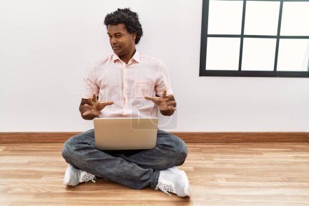 Téléchargez les photos : Homme africain avec les cheveux bouclés à l'aide d'un ordinateur portable assis sur le sol expression dégoûtée, mécontent et peur de faire visage dégoûtant parce que la réaction d'aversion. les mains levées - en image libre de droit