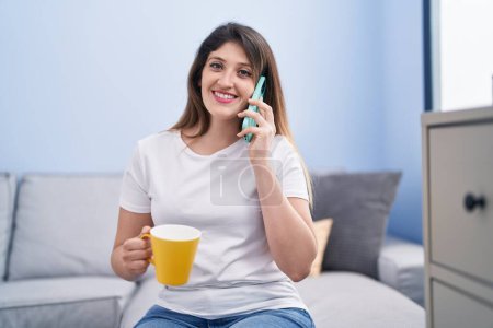 Photo for Young hispanic woman drinking coffee and talking on the smartphone at home - Royalty Free Image