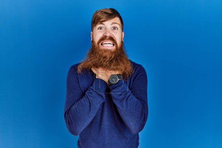 Téléchargez les photos : Homme rousse à longue barbe portant un pull bleu décontracté sur fond bleu criant suffoquer parce que l'étranglement douloureux. problème de santé. concept d'asphyxie et de suicide. - en image libre de droit