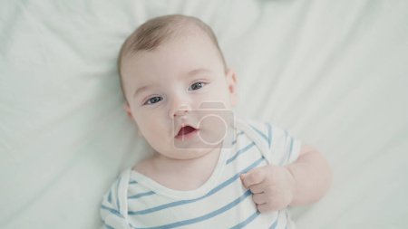 Téléchargez les photos : Adorable bébé caucasien couché sur le lit avec une expression détendue dans la chambre - en image libre de droit