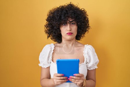 Foto de Young brunette woman with curly hair using touchpad over yellow background skeptic and nervous, frowning upset because of problem. negative person. - Imagen libre de derechos