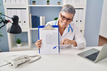 Téléchargez les photos : Femme d'âge moyen portant un uniforme de médecin tenant presse-papiers à la clinique - en image libre de droit