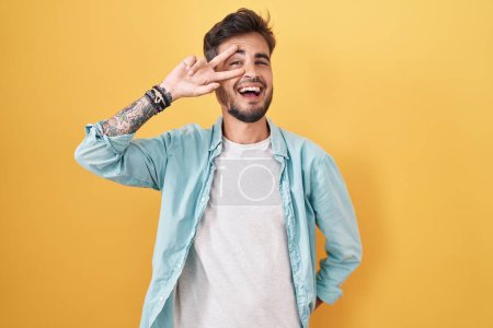 Téléchargez les photos : Jeune homme hispanique avec des tatouages debout sur fond jaune faisant symbole de paix avec les doigts sur le visage, souriant joyeux montrant la victoire - en image libre de droit