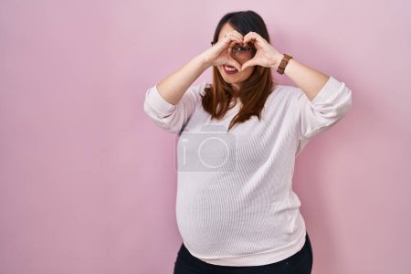 Téléchargez les photos : Pregnant woman standing over pink background doing heart shape with hand and fingers smiling looking through sign - en image libre de droit