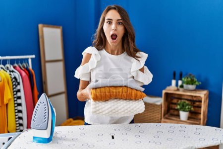 Photo for Young hispanic woman ironing clothes at laundry room holding folded sweaters afraid and shocked with surprise and amazed expression, fear and excited face. - Royalty Free Image
