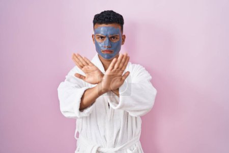 Foto de Joven hombre hispano usando mascarilla de belleza y expresión de rechazo de bata de baño cruzando brazos y palmas haciendo signo negativo, cara enojada - Imagen libre de derechos