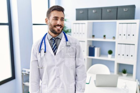 Foto de Hombre hispano guapo vistiendo uniforme médico y estetoscopio en la clínica médica mirando hacia otro lado con sonrisa en la cara, expresión natural. riendo confiado. - Imagen libre de derechos