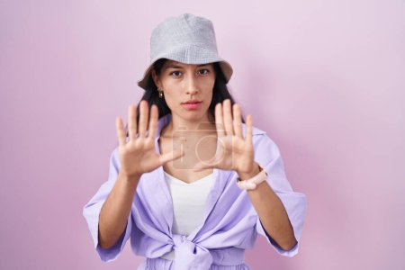 Foto de Mujer hispana joven de pie sobre fondo rosa con sombrero alejando las palmas de las manos mostrando rechazo y negación con miedo y expresión repugnante. parada y prohibido. - Imagen libre de derechos