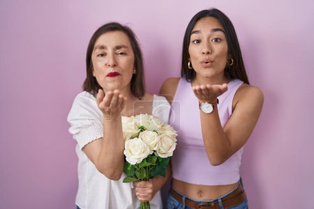 Téléchargez les photos : Mère et fille hispaniques tenant bouquet de fleurs blanches regardant la caméra souffler un baiser avec la main sur l'air étant belle et sexy. expression d'amour. - en image libre de droit