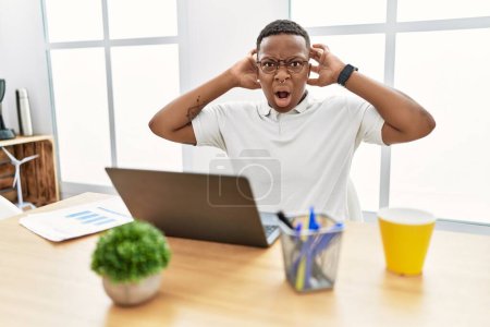 Téléchargez les photos : Jeune homme africain travaillant au bureau en utilisant un ordinateur portable fou et effrayé avec les mains sur la tête, effrayé et surpris de choc avec la bouche ouverte - en image libre de droit