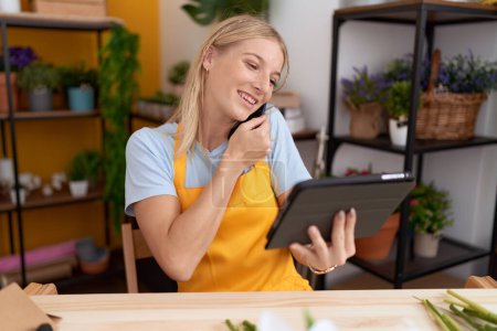 Photo for Young blonde woman florist talking on smartphone using touchpad at flower shop - Royalty Free Image