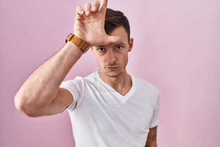 Photo for Caucasian man standing over pink background making fun of people with fingers on forehead doing loser gesture mocking and insulting. - Royalty Free Image