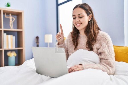 Photo for Young hispanic woman using computer laptop on the bed smiling with an idea or question pointing finger with happy face, number one - Royalty Free Image