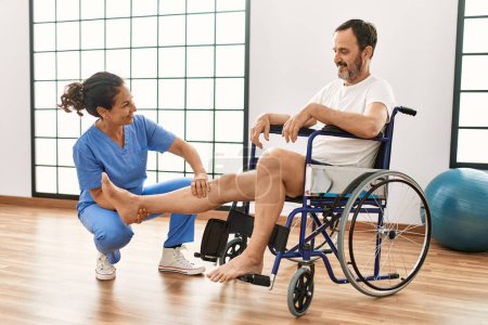 Photo for Middle age man and woman having rehab session manipulating legs sitting on wheelchair at physiotherapy clinic - Royalty Free Image