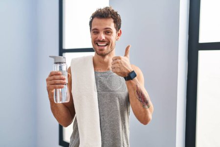 Foto de Un joven hispano usando ropa deportiva bebiendo agua haciendo un gesto feliz con la mano. aprobando la expresión mirando a la cámara mostrando éxito. - Imagen libre de derechos