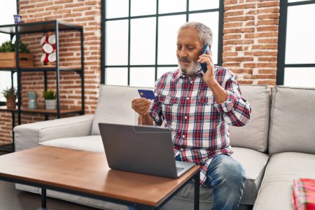 Téléchargez les photos : Homme âgé aux cheveux gris parlant sur un smartphone tenant une carte de crédit assis sur un canapé à la maison - en image libre de droit