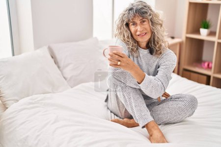 Foto de Mujer de mediana edad bebiendo taza de café sentado en la cama en el dormitorio - Imagen libre de derechos