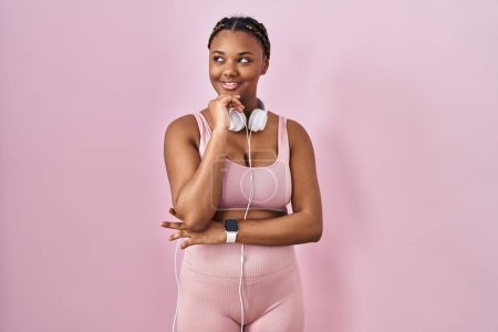 Photo for African american woman with braids wearing sportswear and headphones with hand on chin thinking about question, pensive expression. smiling and thoughtful face. doubt concept. - Royalty Free Image