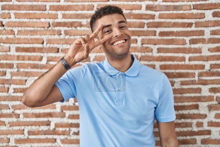 Photo for Brazilian young man standing over brick wall doing peace symbol with fingers over face, smiling cheerful showing victory - Royalty Free Image